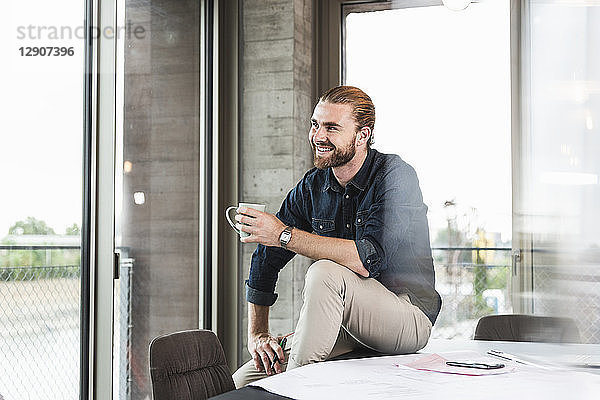 Smiling young businessman sitting on desk in office with cup of coffee