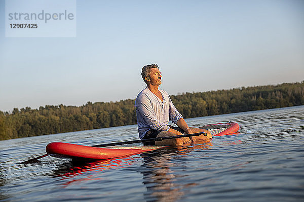 Mature man sitting on paddleboard on a lake by sunset relaxing