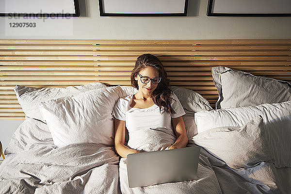 Portrait of smiling young woman lying on bed using laptop