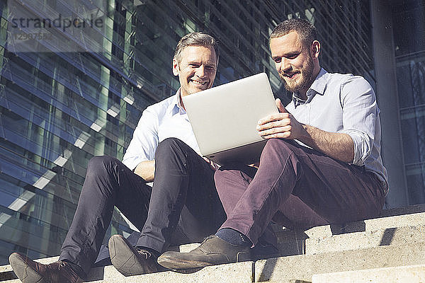 Two smiling businessmen sitting on stairs using laptop