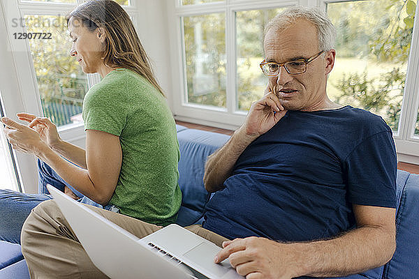 Mature couple sitting on couch at home with man using laptop and woman using cell phone