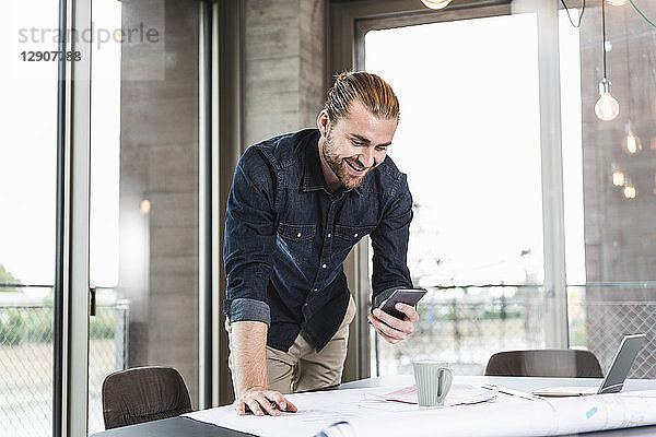 Smiling young businessman at desk in office with plan and cell phone