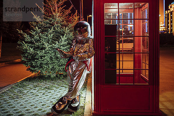 Spaceman leaning against a telephone box at night reading phone book