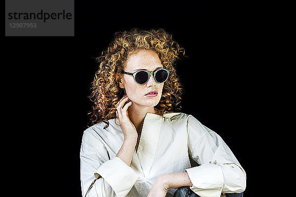 Portrait of stylish young woman with curly red hair wearing sunglasses in front of black background
