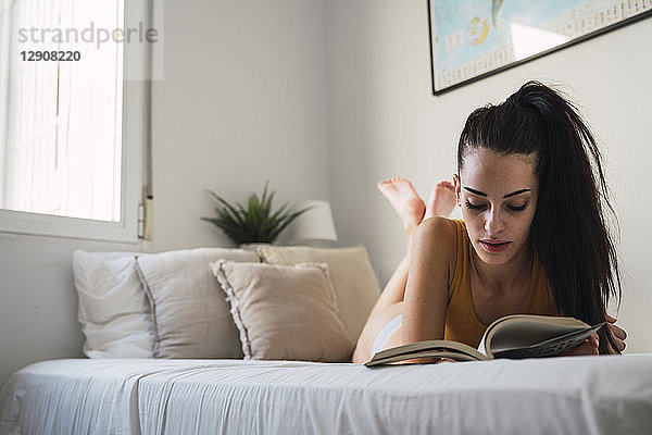 Beautiful young woman lying on bed at home reading a book