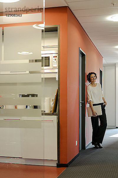 Laughing businesswoman standing in office corridor  holding laptop