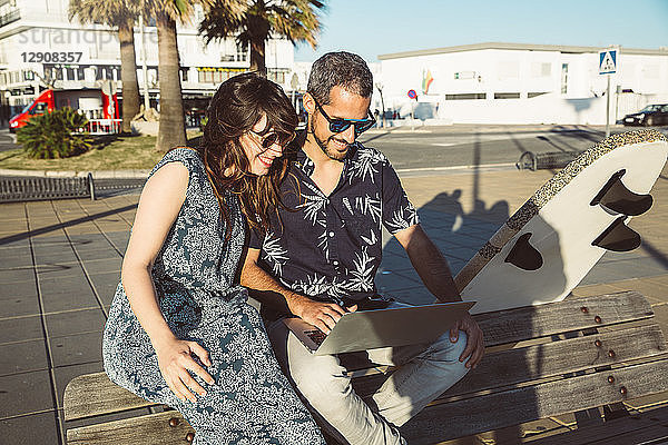 Couple with surfboard sitting on the beach  using laptop