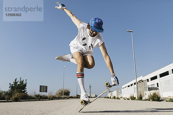 Man in stylish sportive outfit standing on skateboard against blue sky