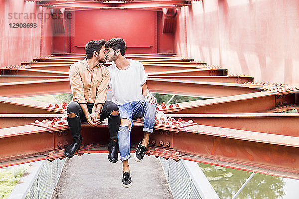 Kissing young gay couple sitting on steel girder of a footbridge