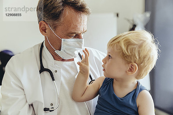 Toddler sitting on lap of pediatrician  wearing protective mask