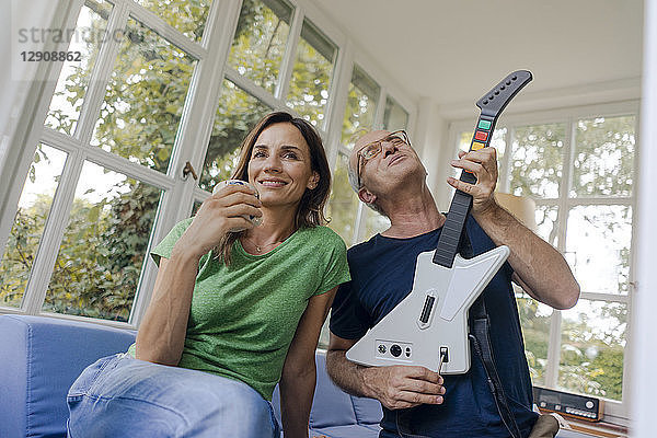 Mature couple sitting on couch at home with man playing toy electric guitar