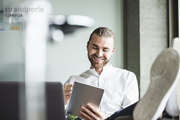 Smiling businessman sitting in office with feet up using tablet