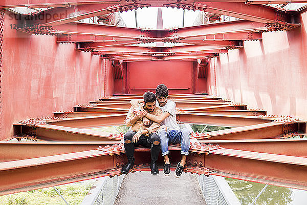 Young gay couple in love sitting on steel girder of a footbridge looking down
