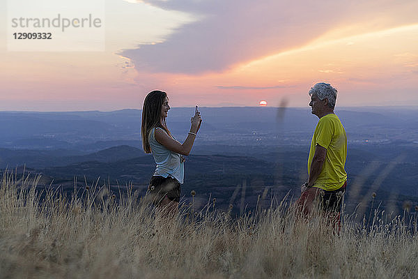 Spain  Catalonia  Montcau  senior father and adult daughter taking a cell phone picture on top of hill during sunset