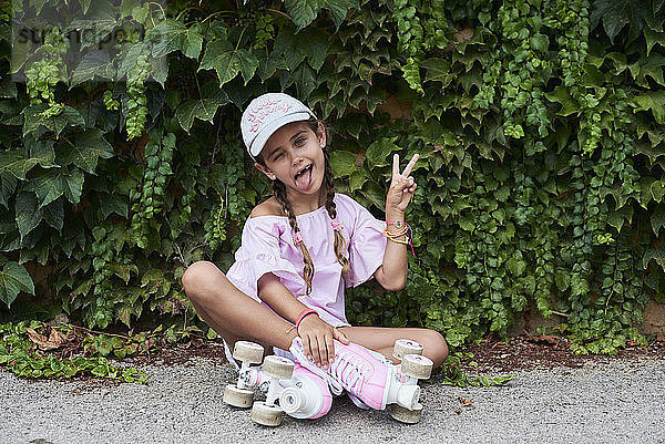 Confident little girl sticking out tongue sitting on the ground with pink roller skates