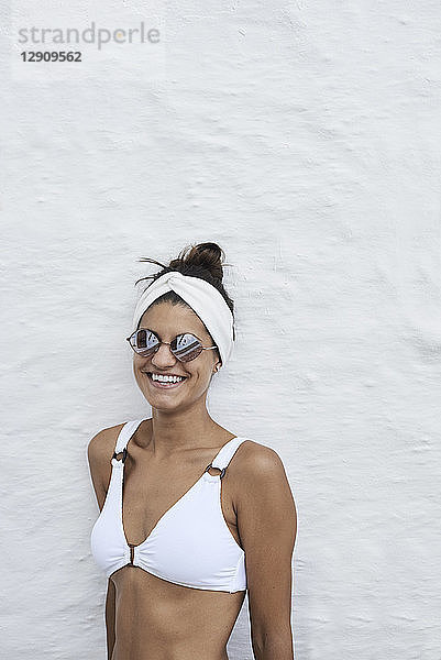 Portrait of laughing young woman wearing white bikini top leaning against white wall