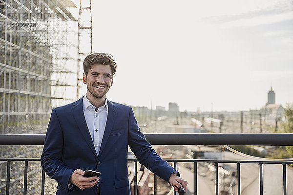 Portrait of smiling businessman standing on bridge in the city holding cell phone