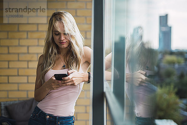 Blond young woman checking cell phone at the window