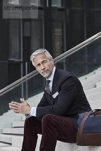 Portrait of fashionable businessman with travelling bag sitting on stairs outdoors