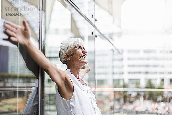 Happy senior woman leaning against glass facade with outstretched arms