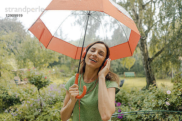 Happy mature woman standing in rain under umbrella listening to music with headphones