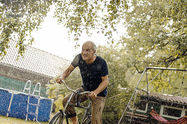 Happy mature man riding bicycle in summer rain in garden