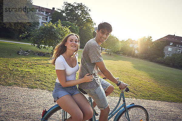 Young couple riding bicycle in park  woman sitting on rack