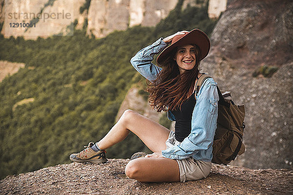 Smiling young woman on a hiking trip wearing a hat sitting on a rock