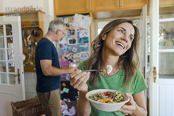 Happy mature woman at home eating a salad with man in background