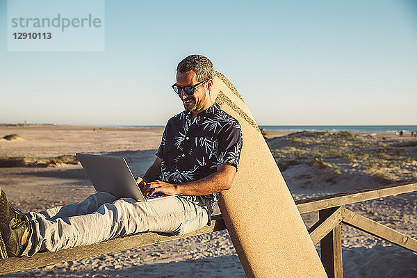 Man sitting at the beach  using laptop  with surfboard leaning on fence