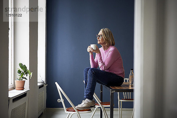 Mature woman at home with cup of coffee looking out of window