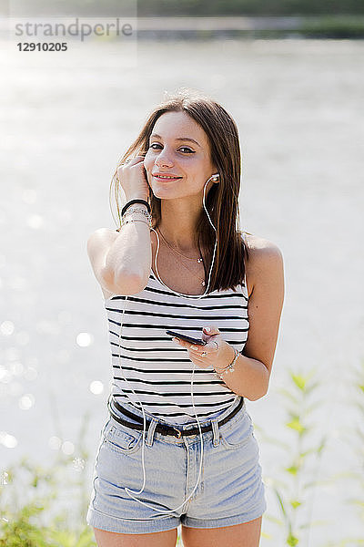 Portrait of smiling young woman listening music with earphones and smartphone at the waterfront