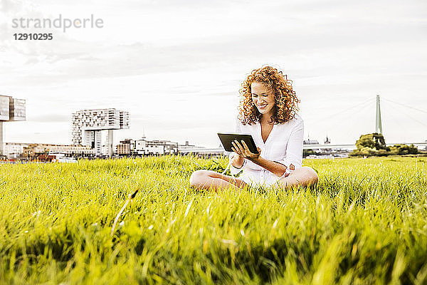 Germany  Cologne  young woman sitting on meadow looking at tablet