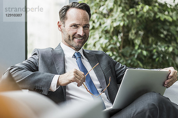 Portrait of smiling businessman sitting in lobby with laptop