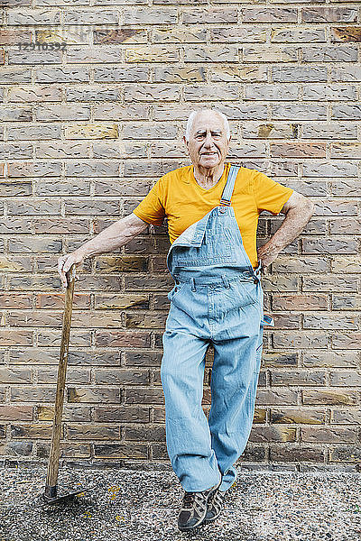 Senior man in denim overall with garden tool looking at camera leaning on a wall