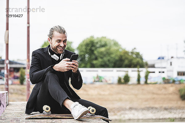Smiling fashionable young man sitting outdoors with cell phone and skateboard
