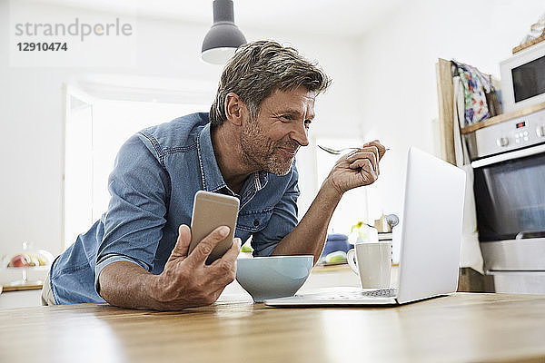 Mature man in his kitchen having breakfast  while checking his laptop