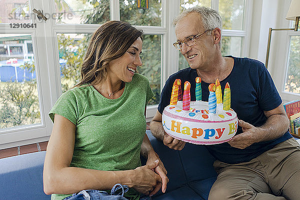 Happy mature couple sitting on couch at home with fake birthday cake