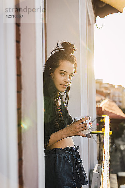 Portrait of beautiful young woman on balcony above the city at sunset with cup of tea