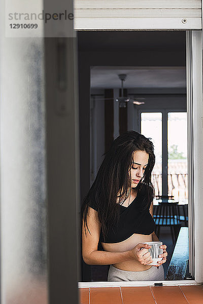 Serious beautiful young woman with cup of coffee at the window at home