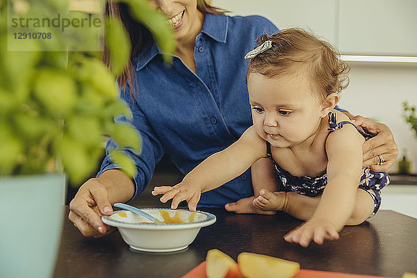 Mother with baby daughter eating fruit pulp in kitchen