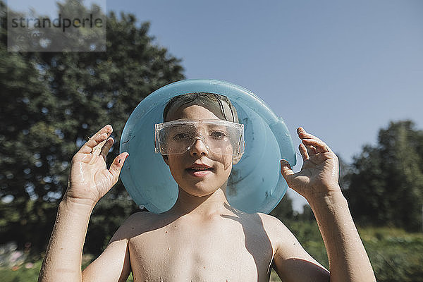 Wet boy wearing safety goggles holding bowl above his head in garden