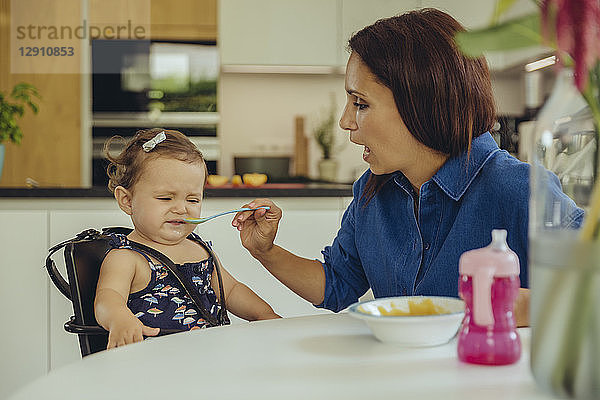 Mother helping disgusted baby daughter eating fruit pulp in kitchen