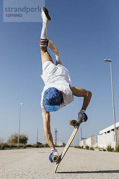 Back view of man in stylish sportive outfit standing on skateboard upside down against blue sky