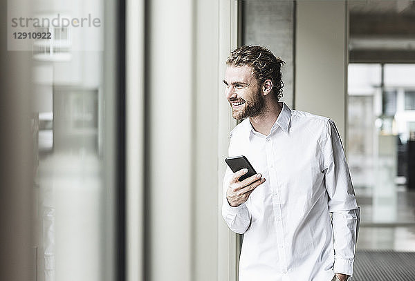 Smiling young businessman holding cell phone looking out of window in office