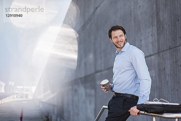 Smiling businessman with laptop bag and takeaway coffee leaning against a railing