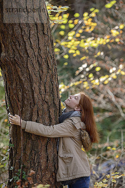 Smiling teenage girl hugging tree trunk in autumnal forest
