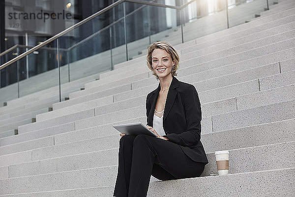 Portrait of smiling businesswoman with tablet and coffee to go sitting on stairs outdoors