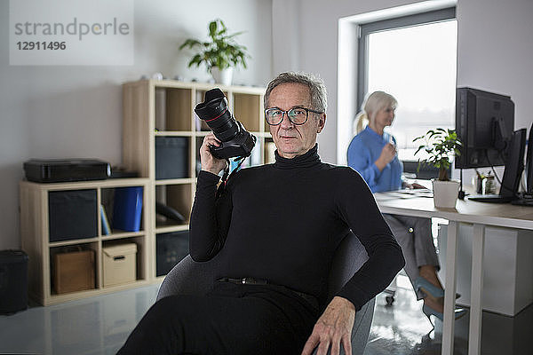 Senior man with camera sitting in office with colleague working behind him