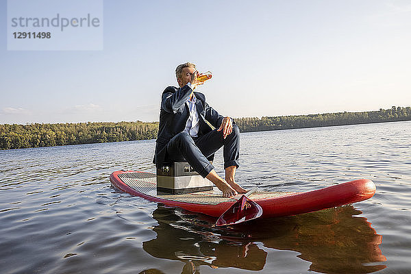 Businessman sitting on surfboard on a lake having a drink